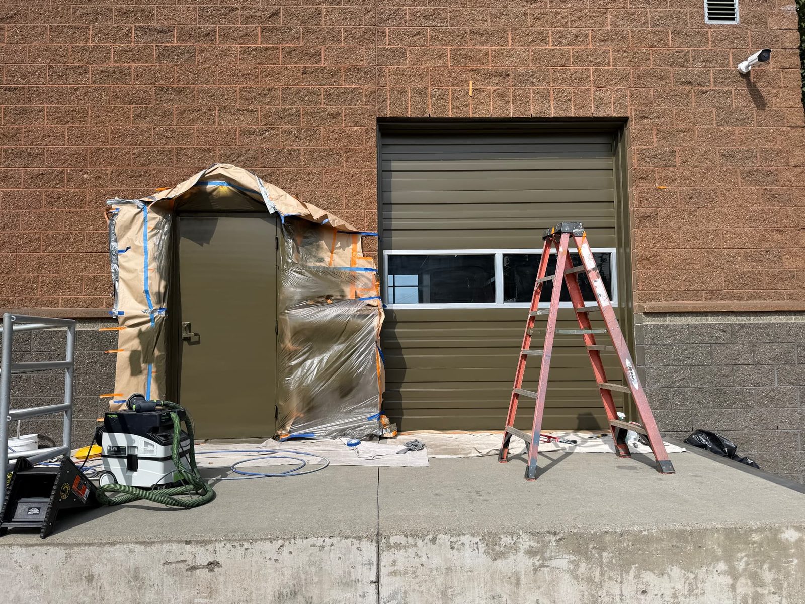 Commercial exterior door masked with paper and plastic sheeting next to a repainted overhead garage door at the Aurora Rents building in Shoreline, with a ladder and painting equipment in place