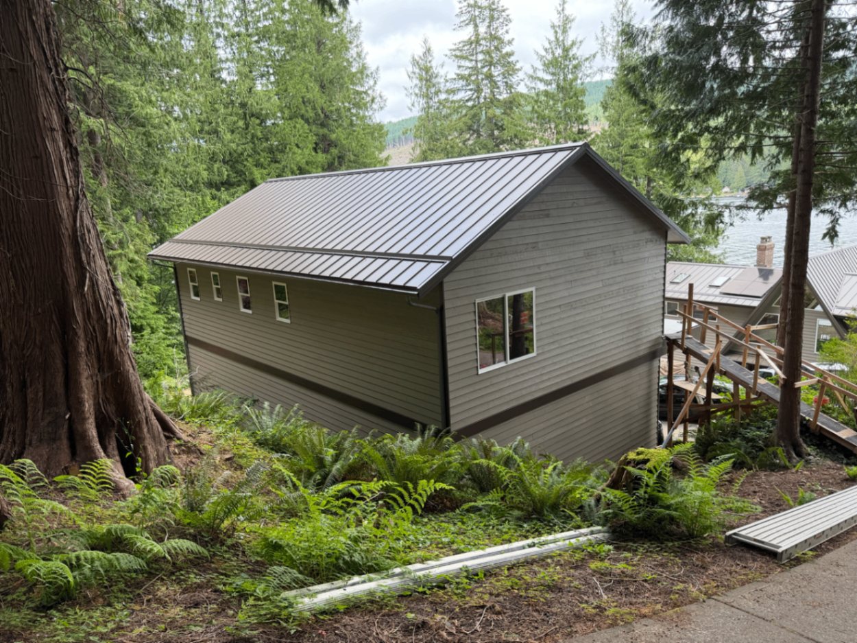 Finished lakefront custom home with olive exterior and dark metal roof set among trees above the lake
